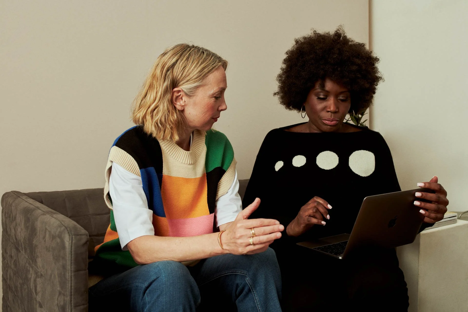 Two women sitting on a sofa looking at a tablet together, one explaining something to the other.