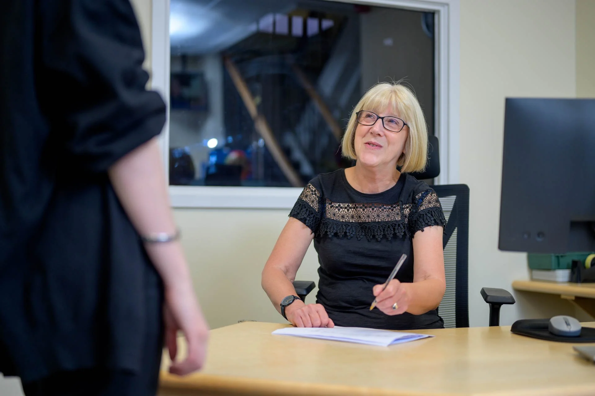 A woman with glasses and blonde hair sitting at an office desk, looking up at a person standing in front of her with a notepad or paper. The woman is smiling and wearing a black top with lace details.