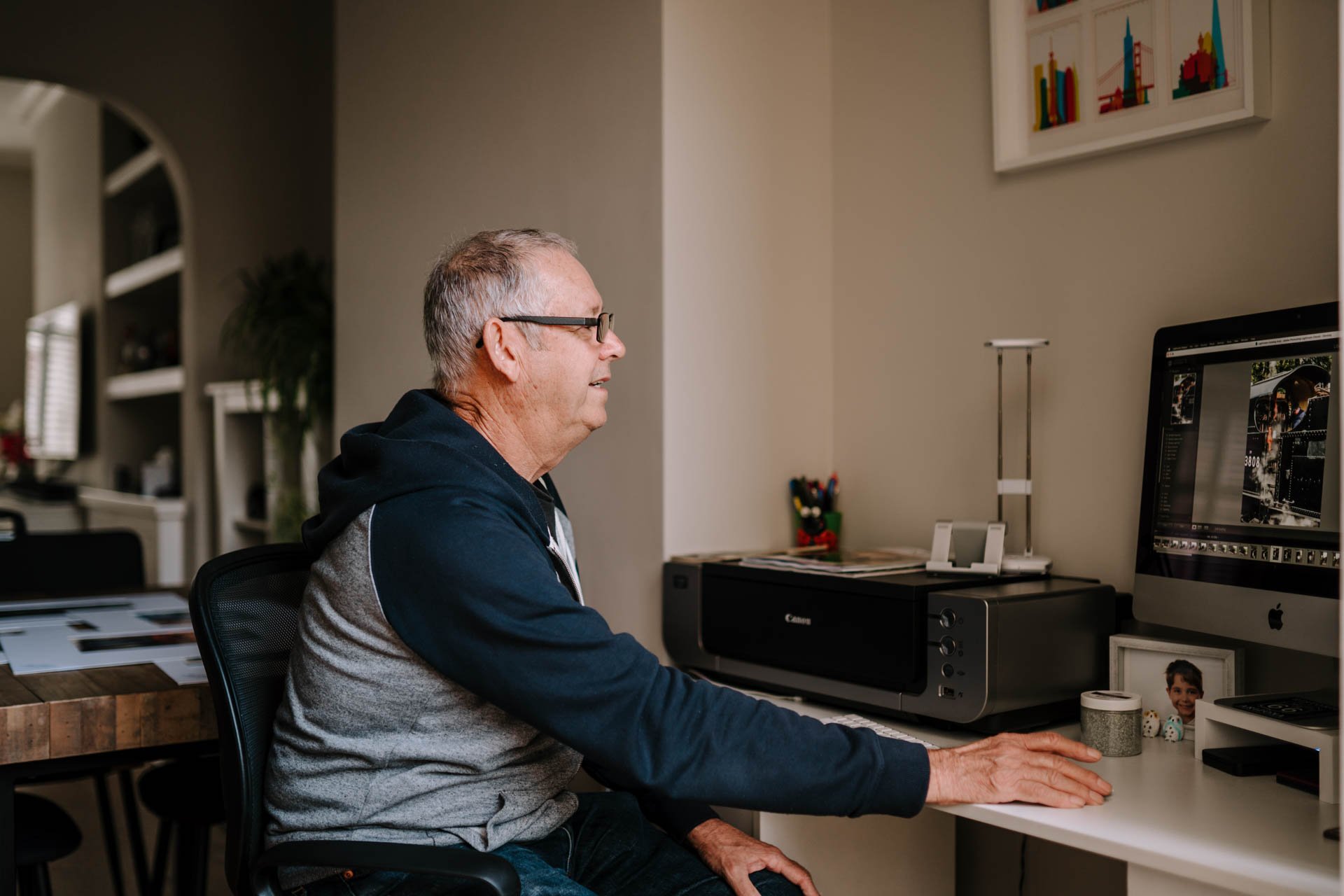 A man with glasses sitting at a desk working on a computer with photo editing software open.