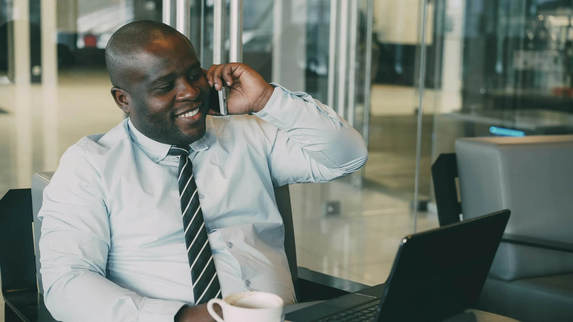 A smiling man in business attire speaking on a mobile phone inside an office, with a laptop and coffee mug on the table.