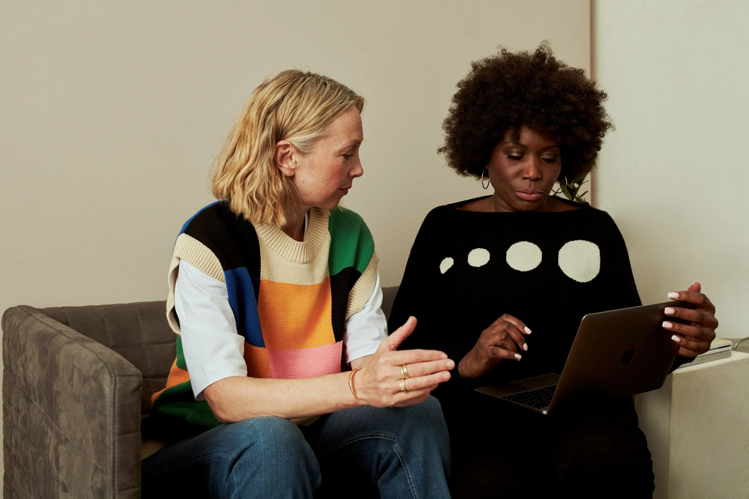 Two middle aged woman one blonde, one black british, dressed in business attire, seated on a sofa, reviewing something on a laptop screen together.