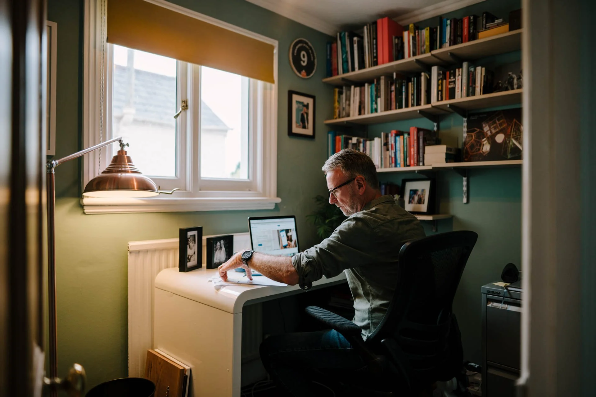 Man sitting at a white desk in a home office, working on a laptop, with bookshelves and framed photos behind him, near a window with a beige shade and a brass desk lamp.