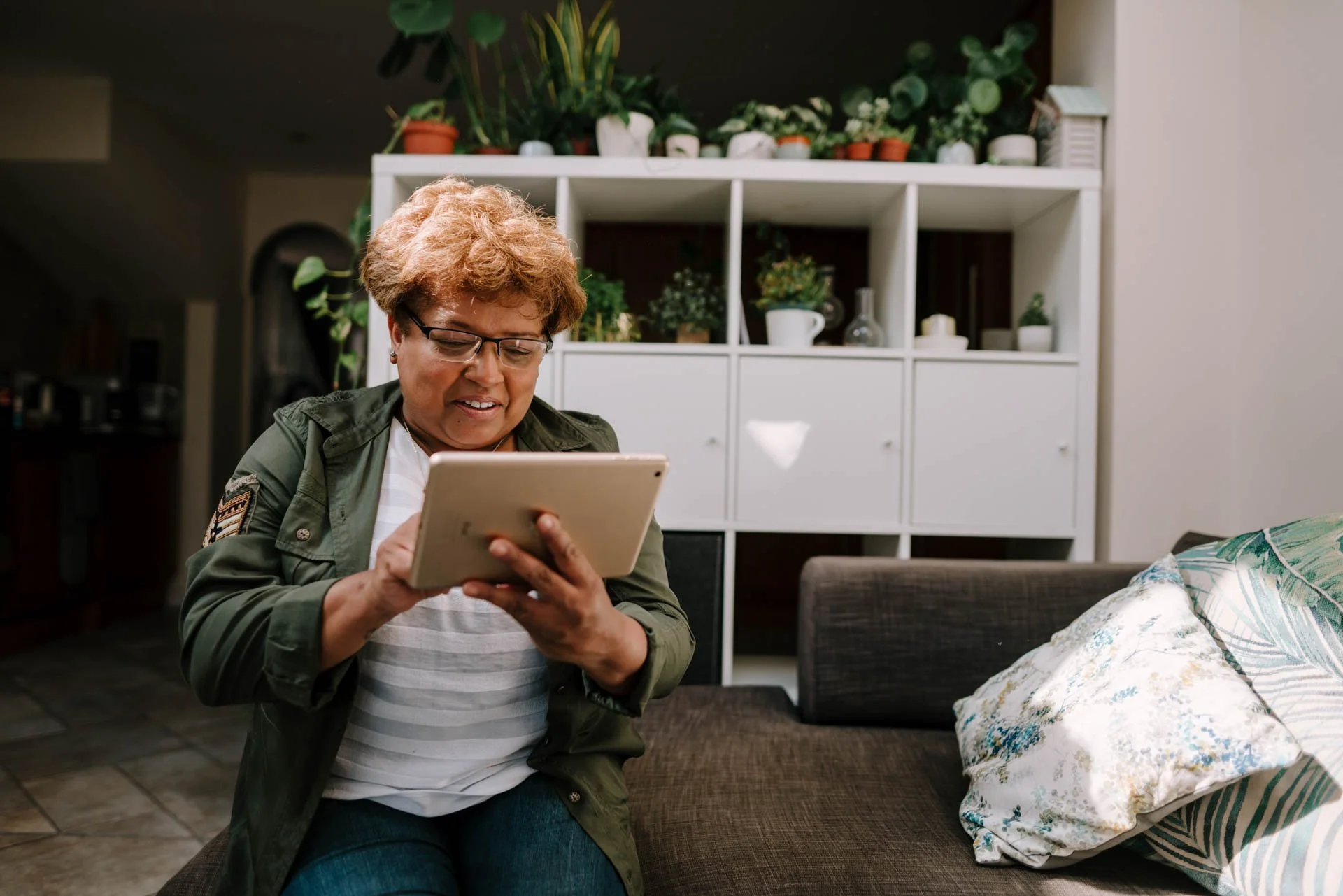 A woman with curly hair wearing glasses, a green jacket, and a white-striped shirt, sitting on a couch, looking at a tablet in her hands in a living room with a white bookshelf with plants and decorative items in the background.
