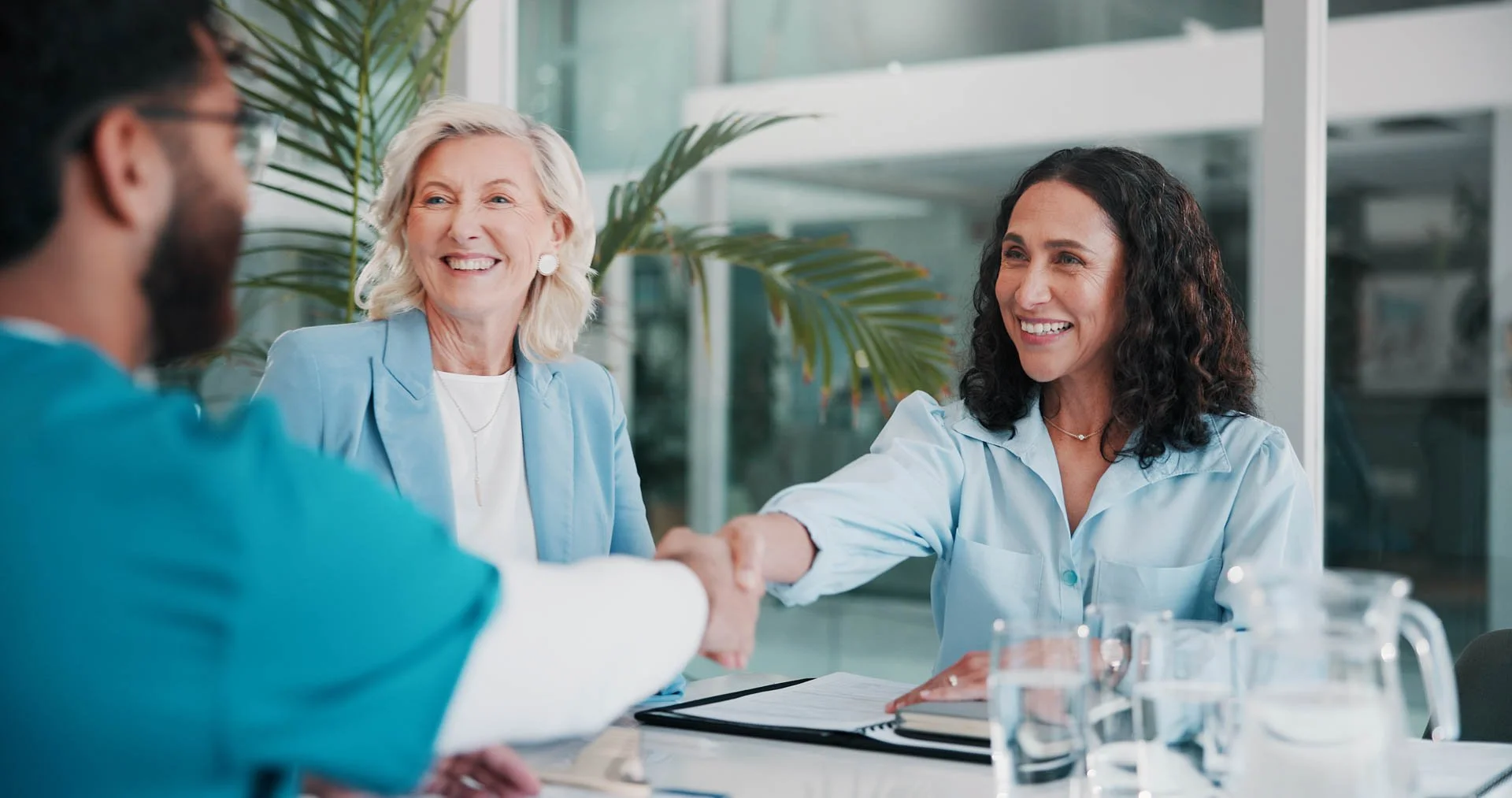 Two women and a man having a meeting, with the women shaking hands, in a bright, modern office with large windows and plants.