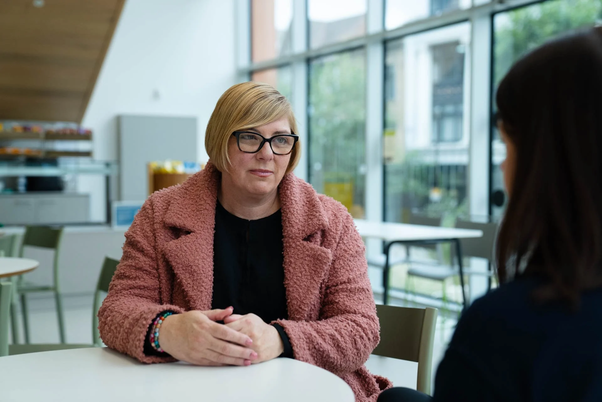 A woman with short blond hair and glasses sitting at a table, listening to another woman with dark hair in a modern, well-lit room with large windows.