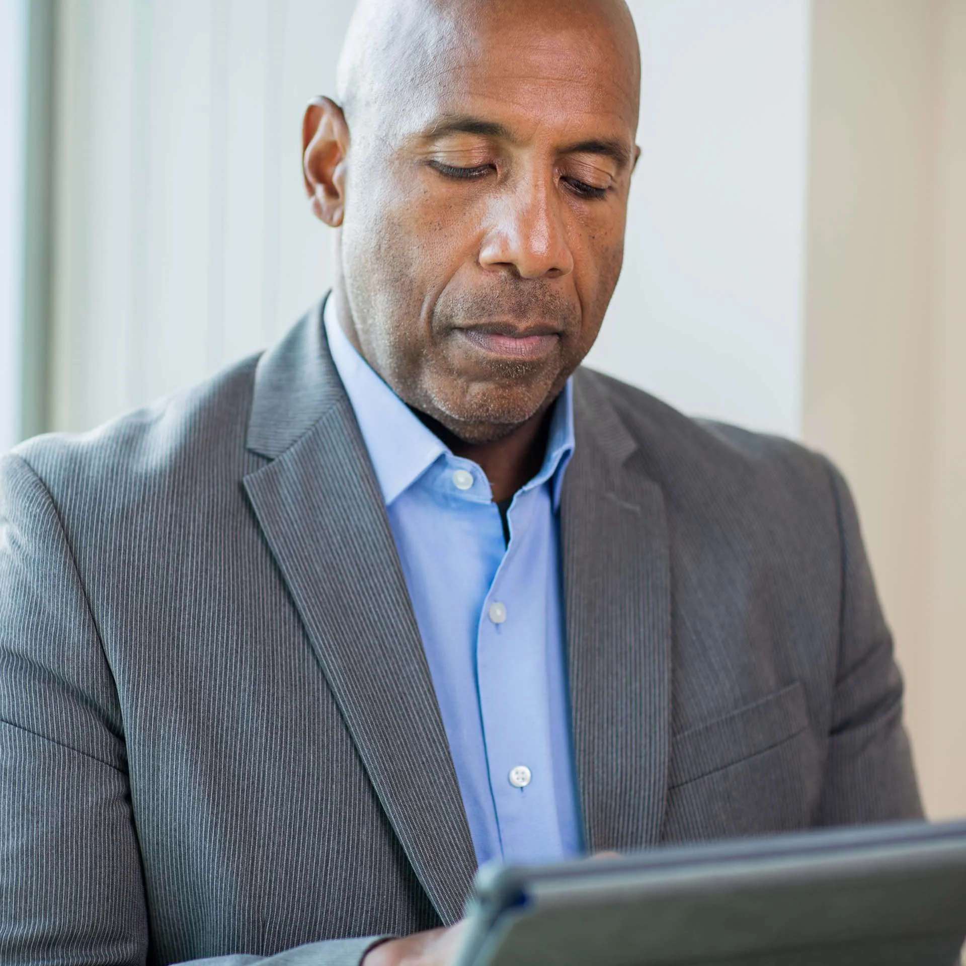 A middle-aged man in a gray business suit with a blue shirt, looking down at a tablet or smartphone.