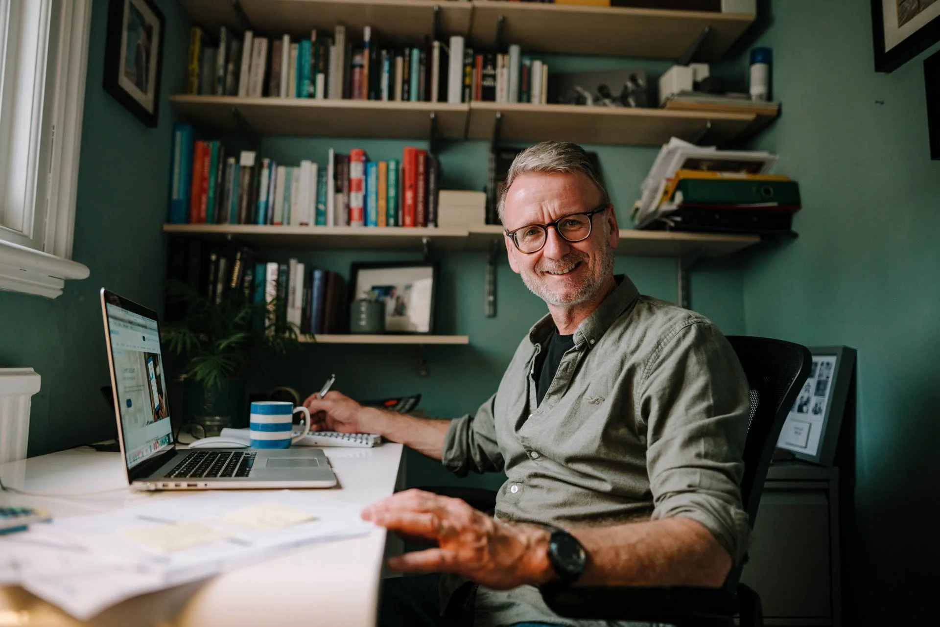 A middle-aged man with glasses and a beard smiling while sitting at a cluttered desk with a laptop, coffee mug, and papers in a home office, with bookshelves and framed pictures in the background.