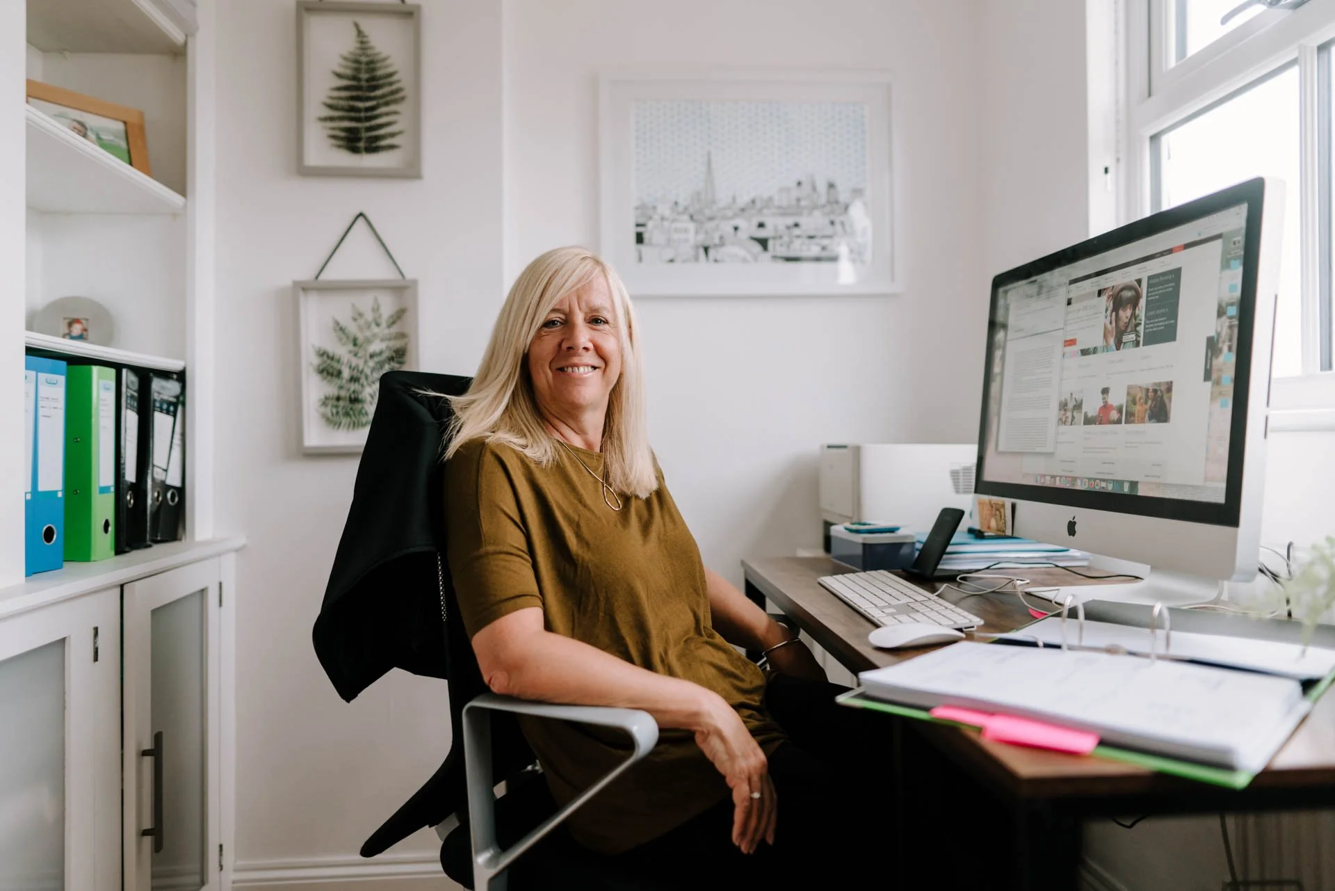 A woman sitting at a desk in a home office, smiling at the camera, with a computer monitor, keyboard, mouse, notebook, and office supplies in the background.