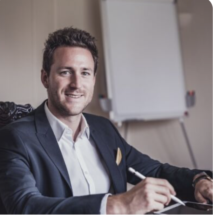 A man in a business suit sitting at a desk with a whiteboard in the background, smiling and holding a pen.