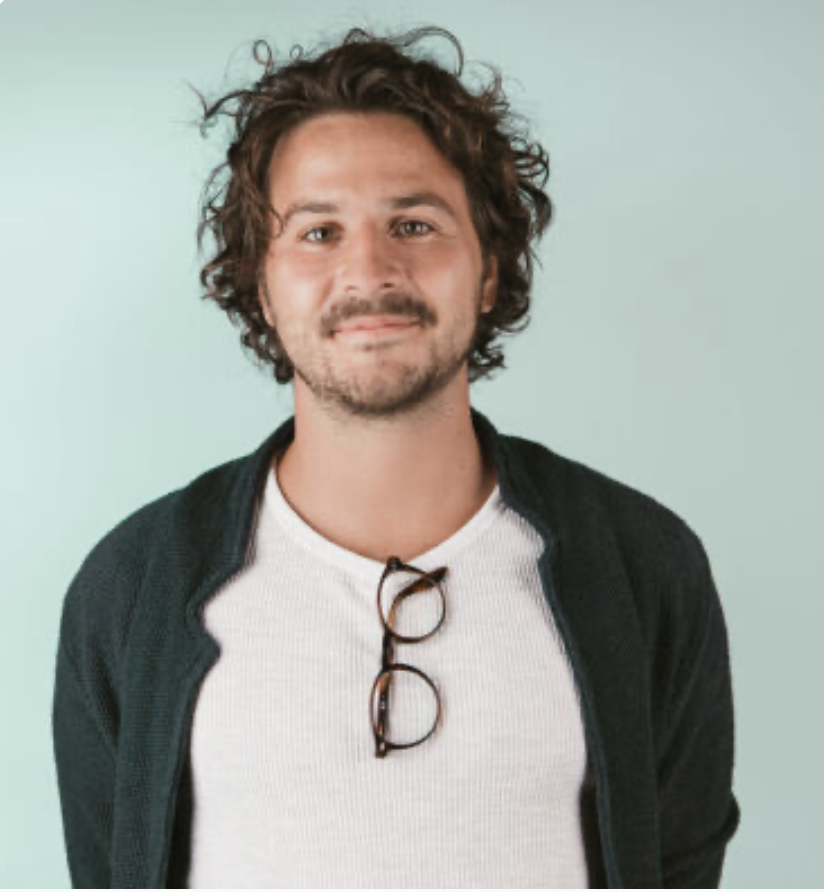 Pascal Brotzer a man with curly hair and a beard, wearing a white t-shirt and a dark jacket, standing against a light green background.