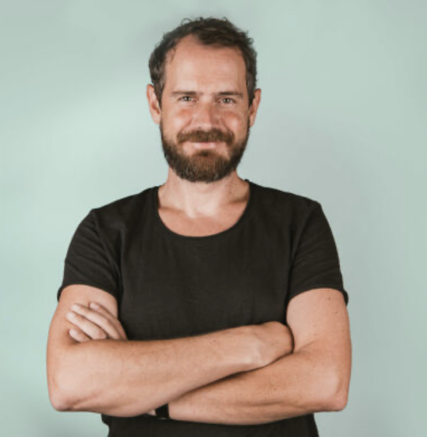 A man with a beard and short hair, wearing a black T-shirt, standing with arms crossed in front of a light green background.