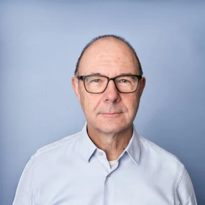 Portrait of an older man named Peter Kaminski with glasses and short hair, wearing a white button-up shirt, against a blue background.