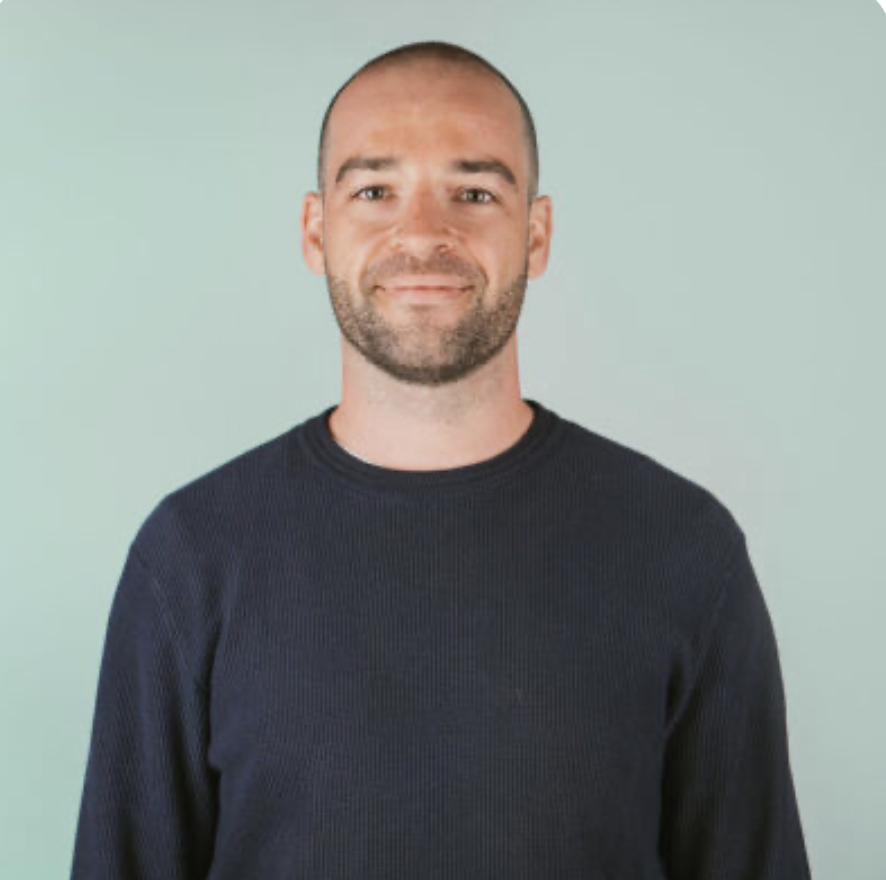 Young man with short hair sitting on a bench in beige pants and a gray polo shirt, against a beige wall, casting a shadow.