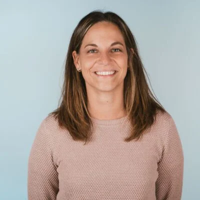 Ashley Stutz, a woman with shoulder-length brown hair smiling, wearing a light pink sweater, in front of a light blue background.