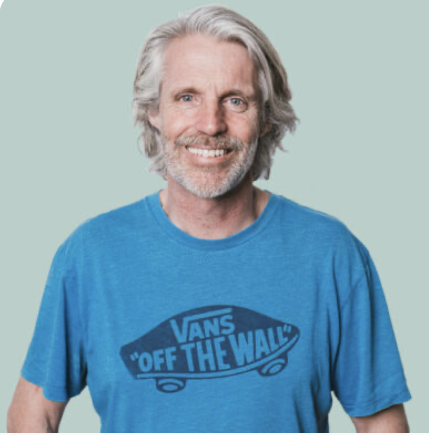 A smiling man with gray hair and beard wearing a blue Vans t-shirt against a light green background.