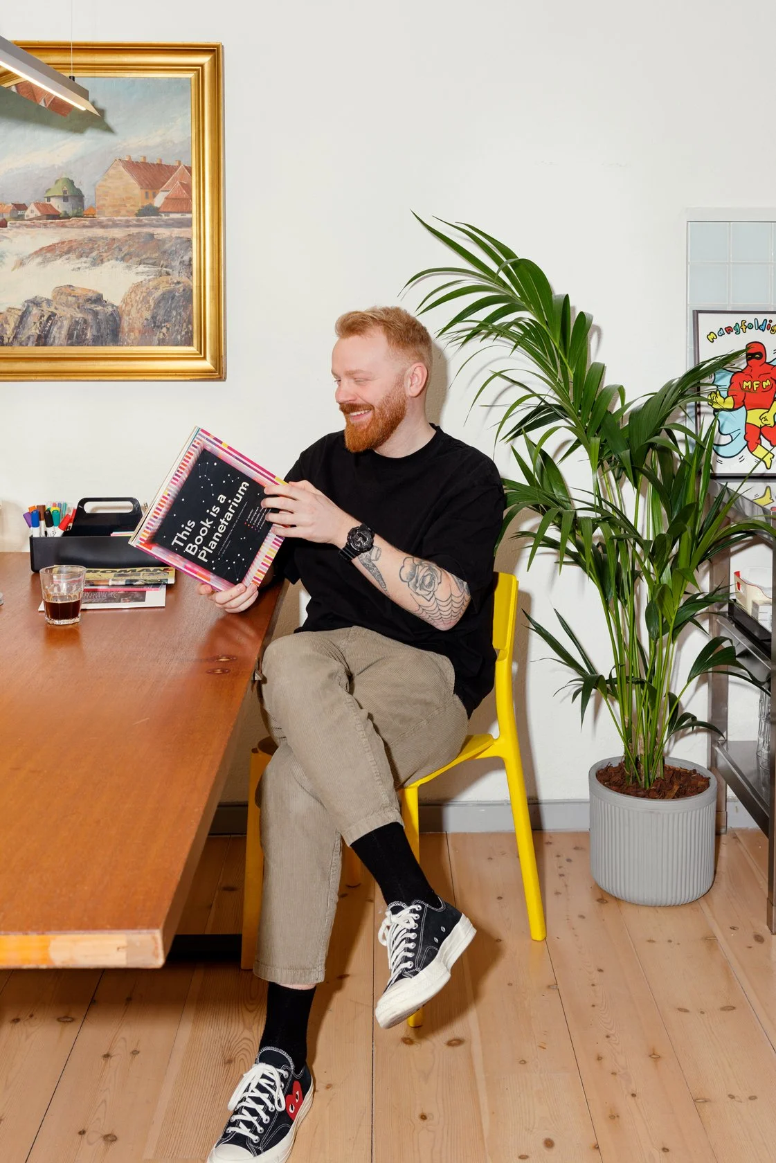 A man with red hair and a beard, wearing a black t-shirt and beige pants, is sitting on a yellow chair at a wooden table, smiling and looking at a book titled "This Book is a Planetarium." The room has a large potted plant, a framed landscape painting, and a colorful poster on the wall.