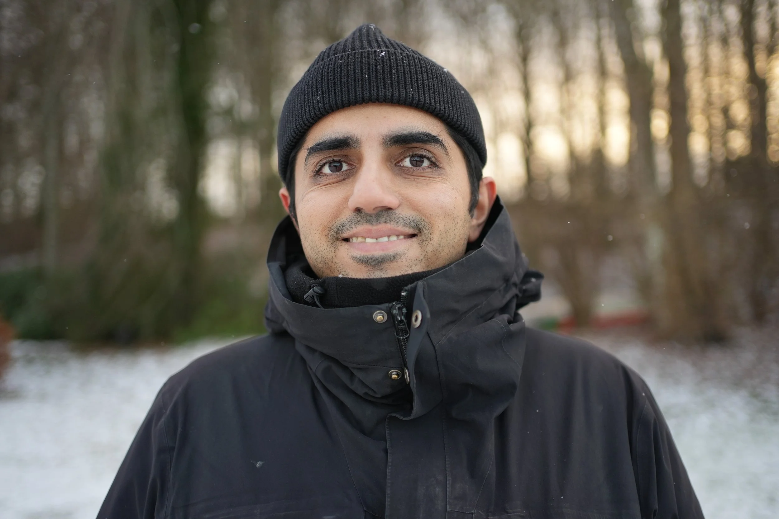 Close-up of a man wearing a black beanie and black winter jacket outdoors with blurred trees and snow in the background.