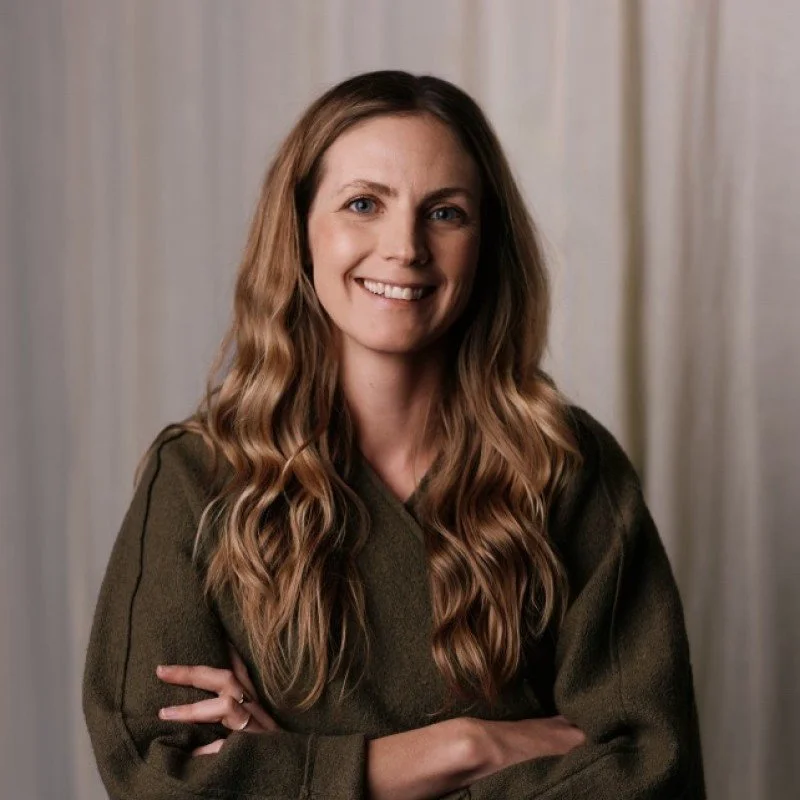 A woman with long wavy brown hair smiling at the camera, wearing a dark green sweater, standing in front of a neutral-colored curtain.