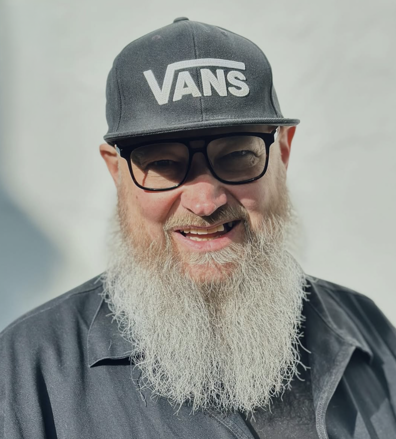 A smiling man with glasses, a gray Vans cap, and a long white beard.