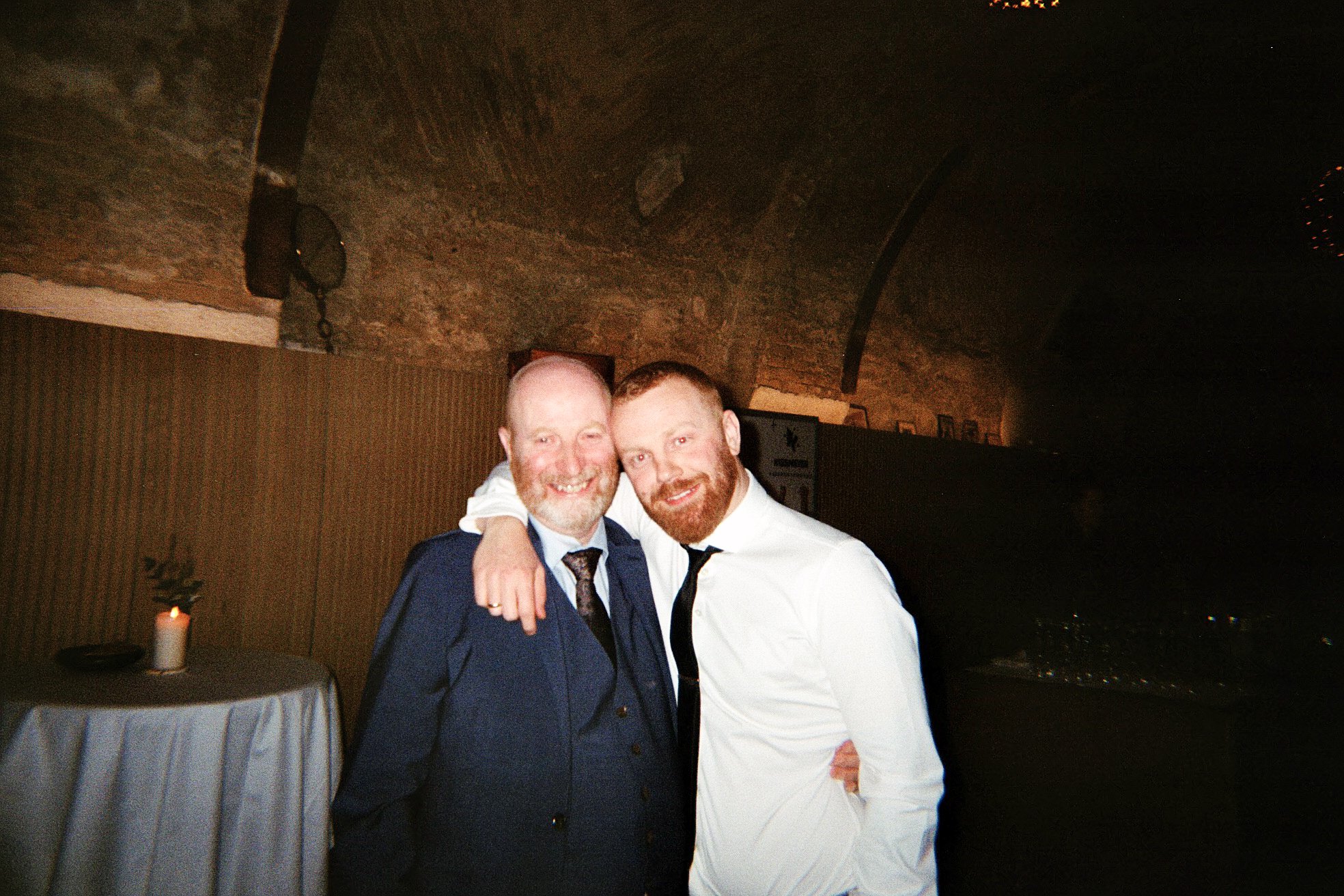 Two men smiling and hugging at a party or celebration, one wearing a dark suit with a tie and the other in a white shirt and black tie, with a candle on a table in the background and a brick arched ceiling.