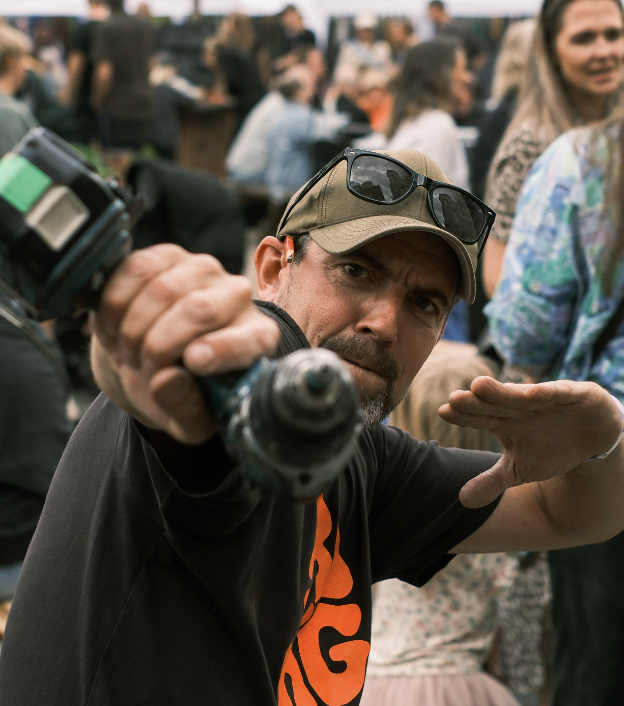 Man wearing a beige cap with sunglasses on top, pointing a camera towards the viewer, with a crowd of people in the background.