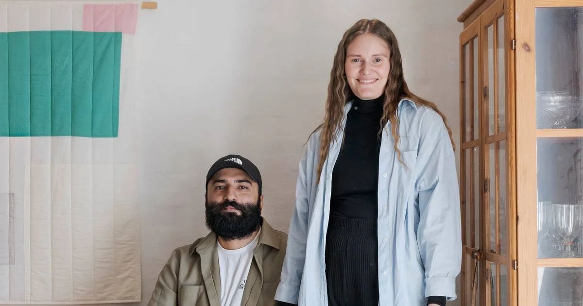 A woman with long wavy hair, wearing a black turtleneck and light blue shirt, smiling at the camera. A man with a beard, wearing a cap and beige jacket, sits beside her.