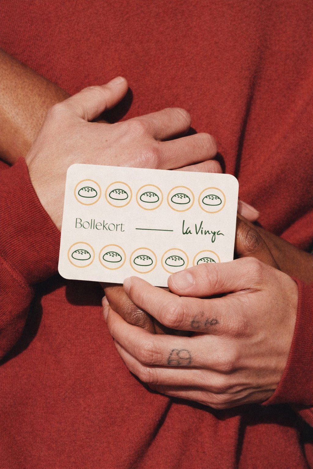 Person holding a card with a pattern of Bread pizza icons, text 'Bollekort LaVinya', and hands with tattoos, wearing a red long-sleeve shirt.