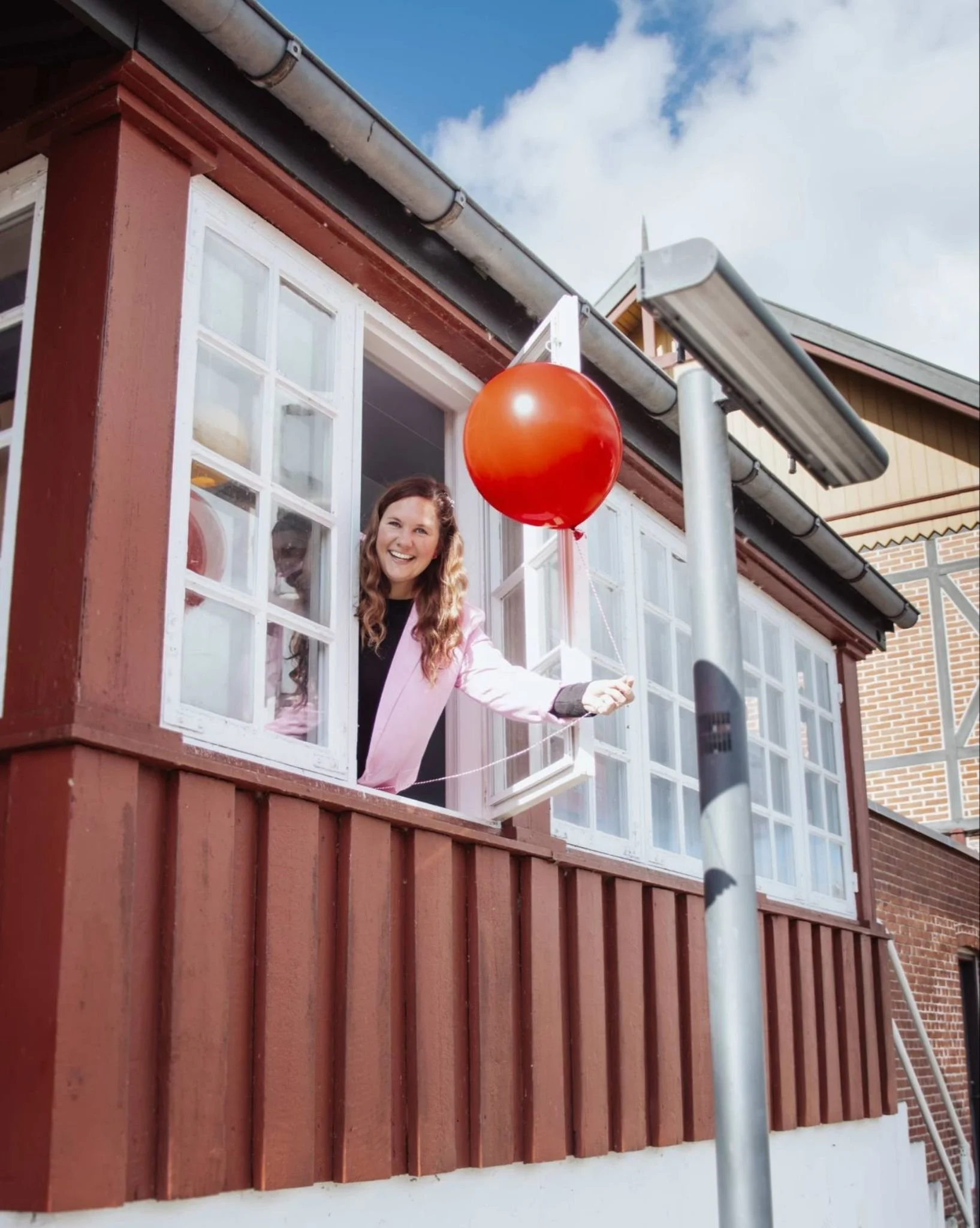 A person with long brown hair and a pink blazer is smiling and standing at a window of a red house with white window panes. There is a red balloon floating in front of the window, and a streetlamp or pole is visible in the foreground. The house has a black roof and a downspout pipe, and the background features a blue sky with white clouds.