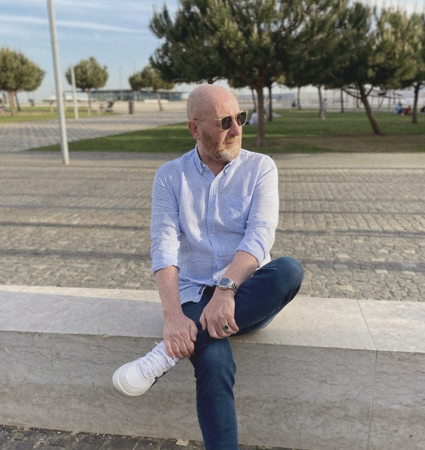 A bald man with a beard wearing sunglasses, a light blue button-up shirt, dark jeans, and white sneakers, sitting on a concrete ledge in a park with trees and a paved walkway in the background.