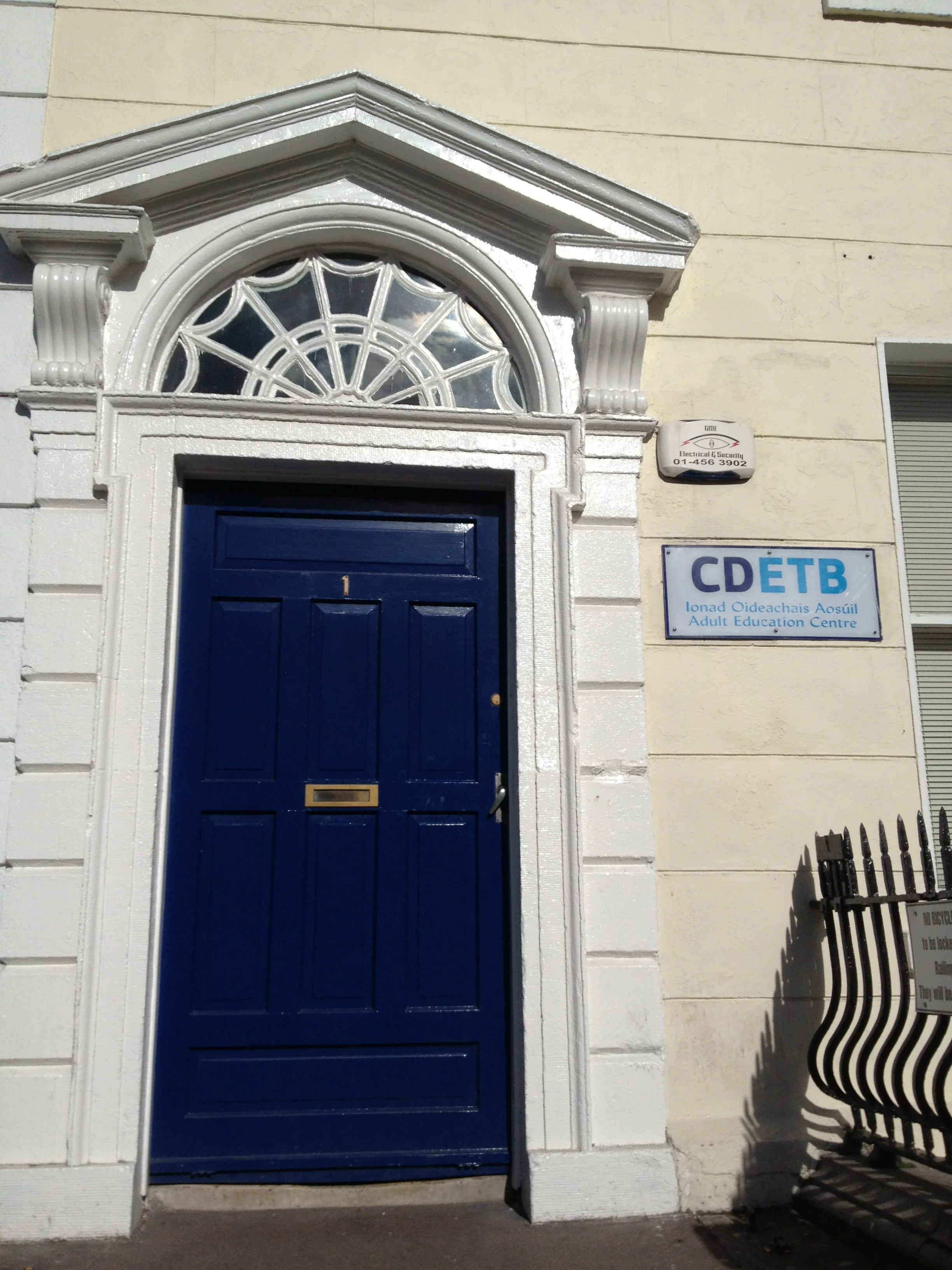 Entrance door to the Adult Education Centre (CDET B) building with a blue door, white decorative archway, and sign indicating the centre's name. Small security sign above and black metal fence nearby.