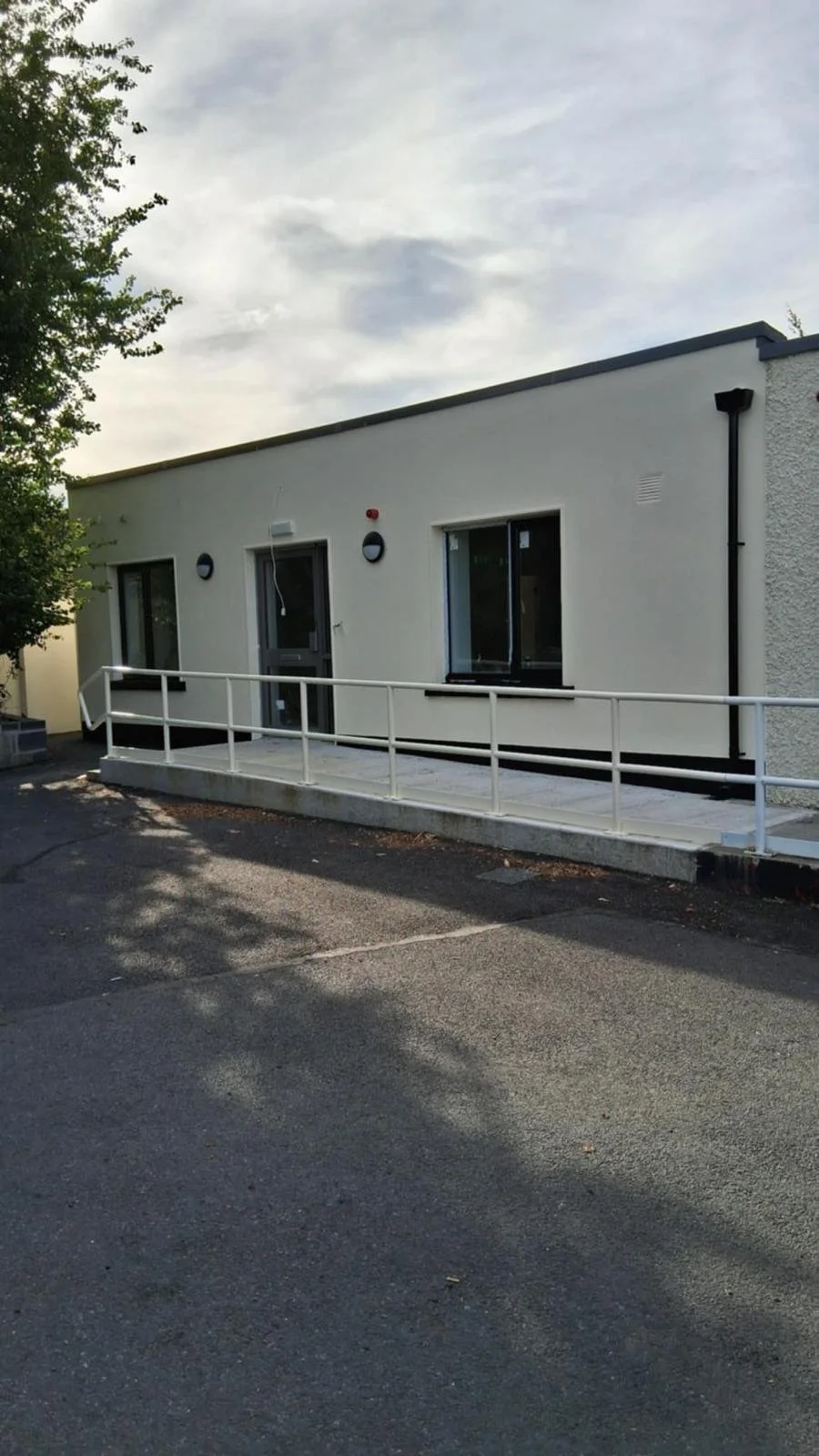 A small, modern building with a wheelchair accessible ramp, white exterior walls, two windows, and a door, outside on an asphalt surface under a cloudy sky.