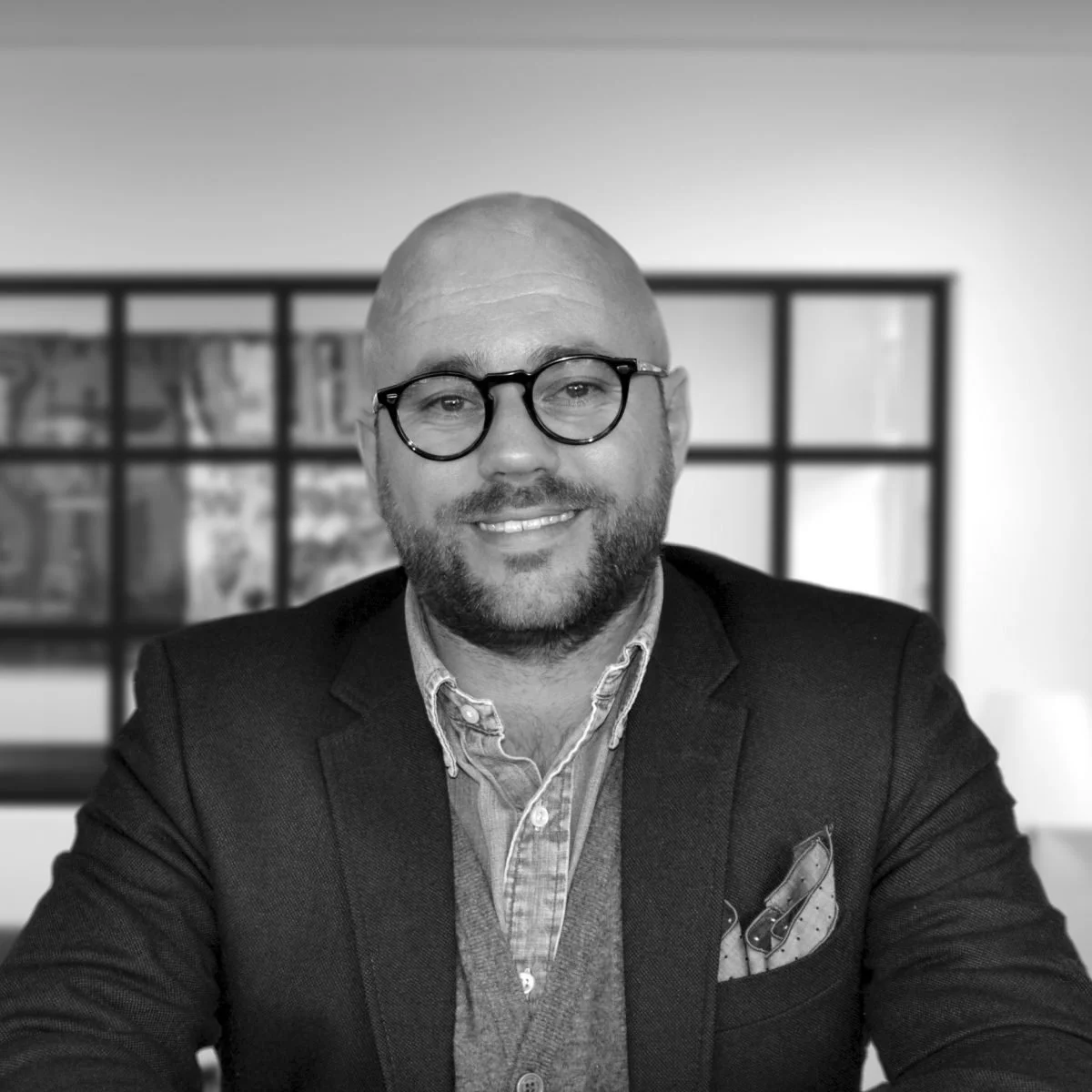A smiling man with a beard and glasses wearing a suit jacket, shirt, and vest, sitting at a table in a room with a large window behind him.