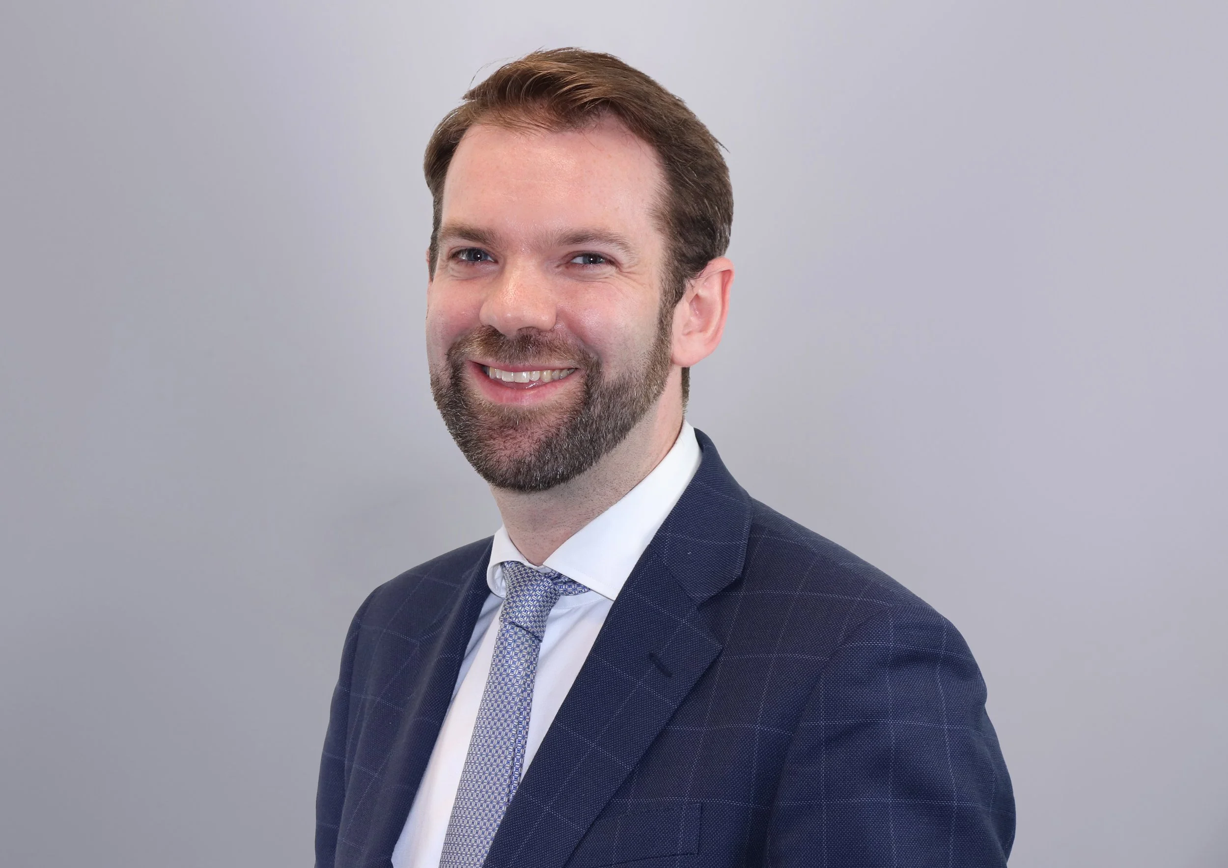A picture of Frederick Way, Mediator.  The picture shows a man with brown hair and beard, wearing a navy blue suit, white shirt, and a patterned tie, smiling against a plain light gray background.