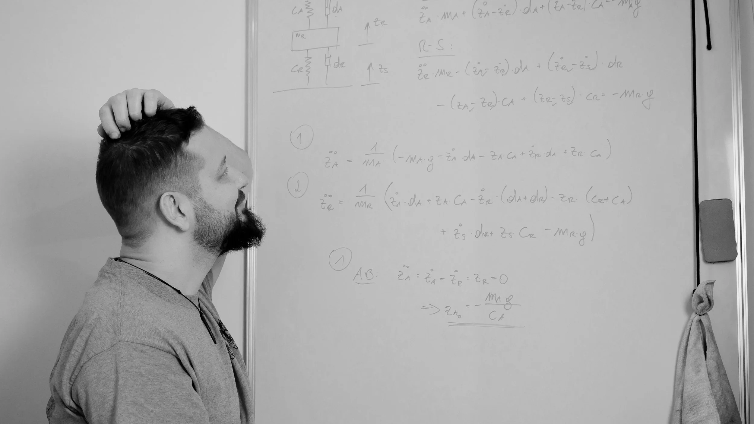 Ingenieur mit Bart und grau-weißem T-Shirt steht vor einem Whiteboard, das mit mathematischen Gleichungen beschrieben ist, und erscheint nachdenklich, die Hand an den Kopf gelegt.