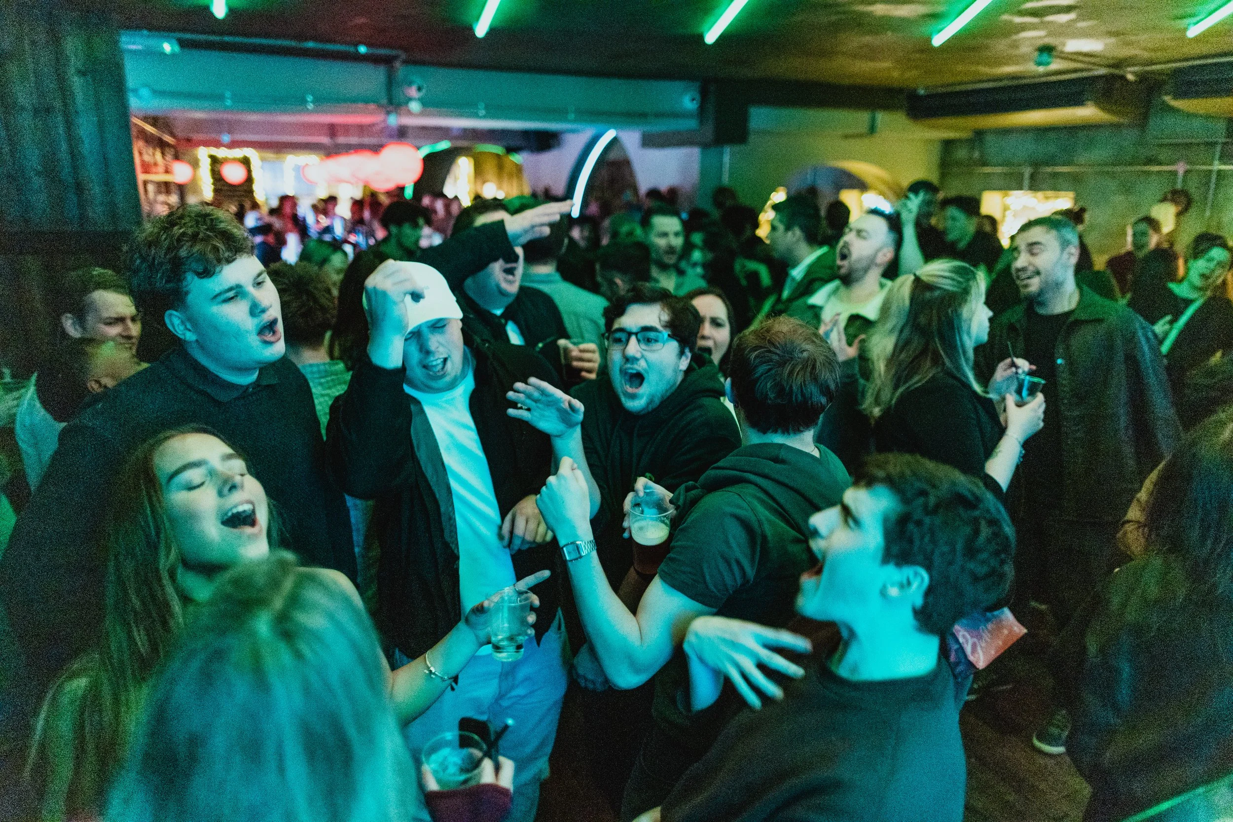 A crowded bar or nightclub with young people dancing, drinking, and socializing under colorful lights, with a lively atmosphere.