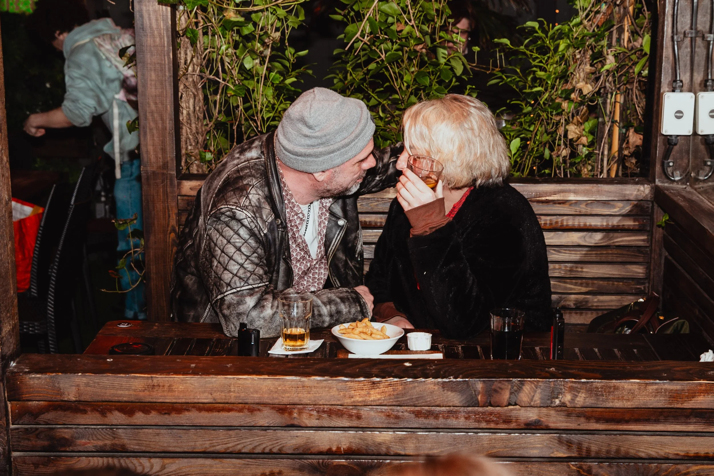 A man and woman are sitting closely at a wooden table in an outdoor restaurant or bar, sharing a moment together. The man is wearing a gray beanie and leather jacket, and the woman has blonde hair and glasses, holding a drink near her mouth. There is