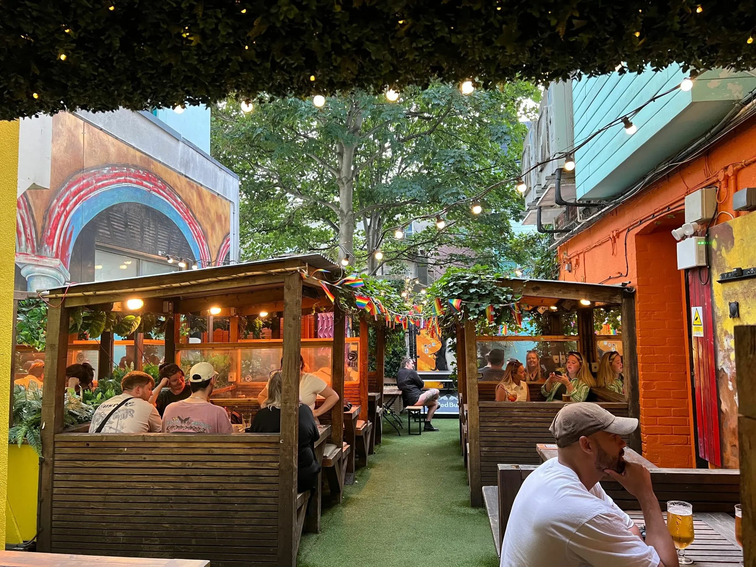 People sitting in outdoor patio of a colorful bar or restaurant, with string lights, greenery, and painted walls, enjoying drinks in the evening.