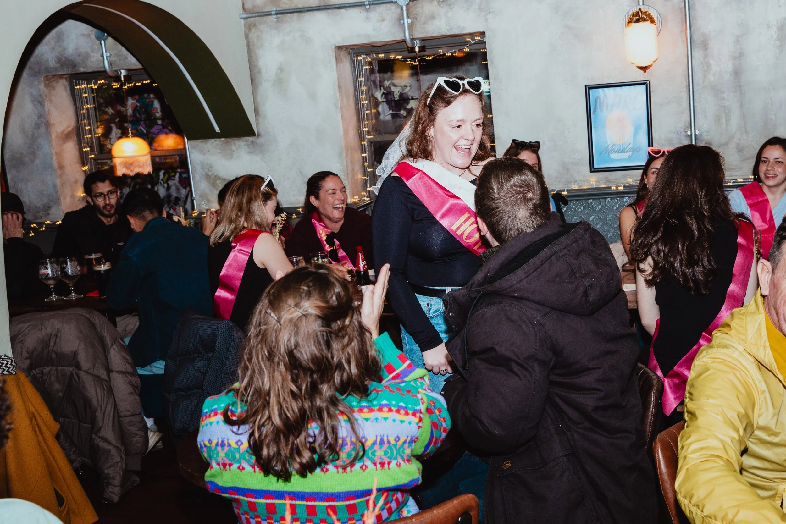A woman with a pink sash and heart-shaped sunglasses smiles and holds hands with a man, while others around them wear pink sashes and enjoy a social gathering in a decorated indoor space with string lights and artwork on the walls.