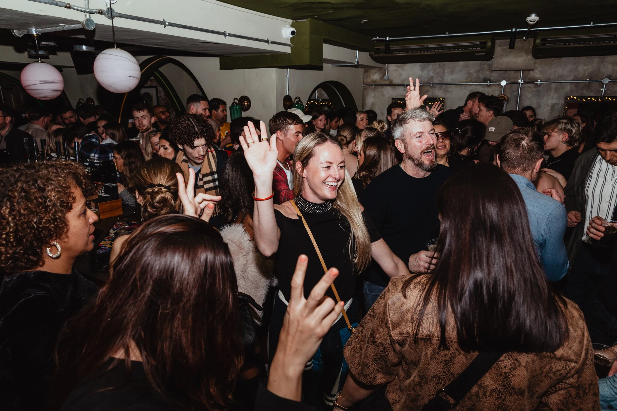 Crowd of people at a lively indoor party or concert, some with raised hands, smiling and enjoying the music.