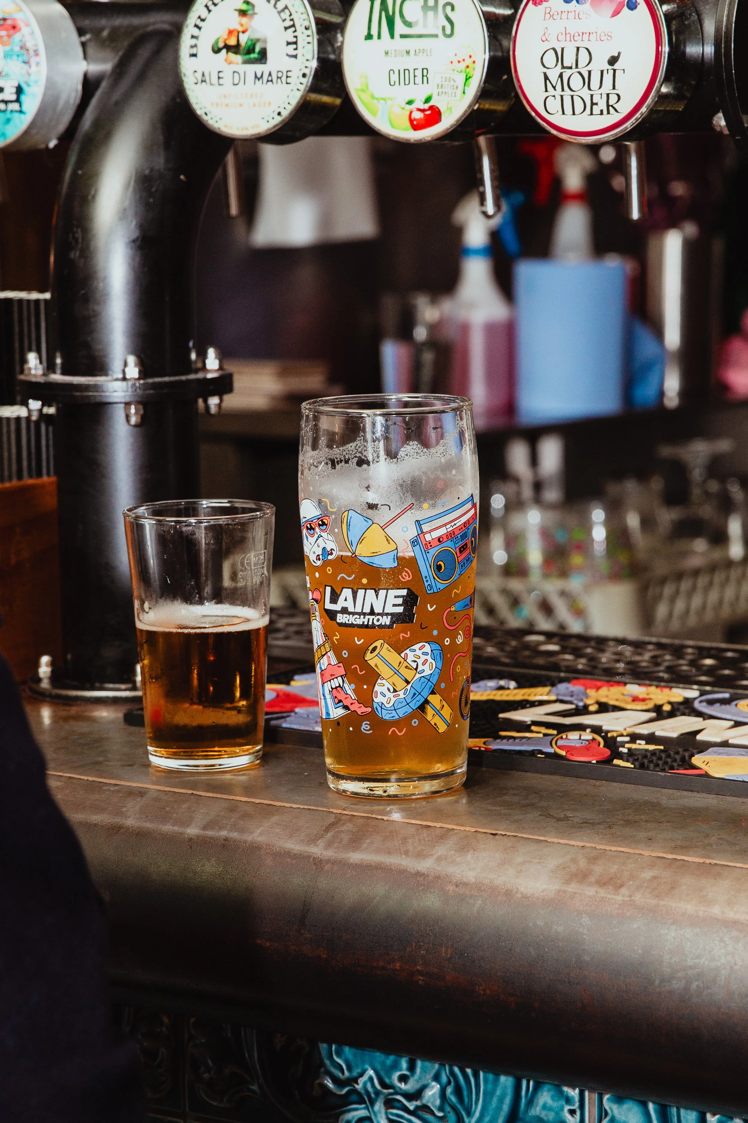 A bar scene with two glasses of beer on a wooden bar counter, one smaller glass, and one larger glass with a colorful LAINE Brighton logo and illustrations. In the background, there are beer taps with various cider labels, and behind the counter, the
