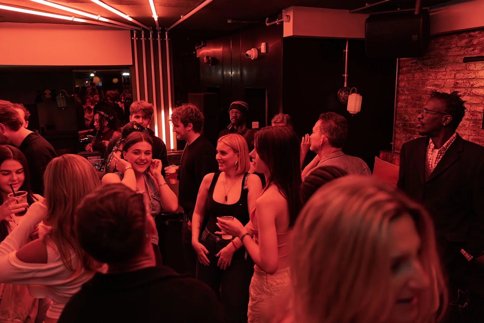 People socializing and dancing at a party in a dimly lit venue with red lighting and brick walls.