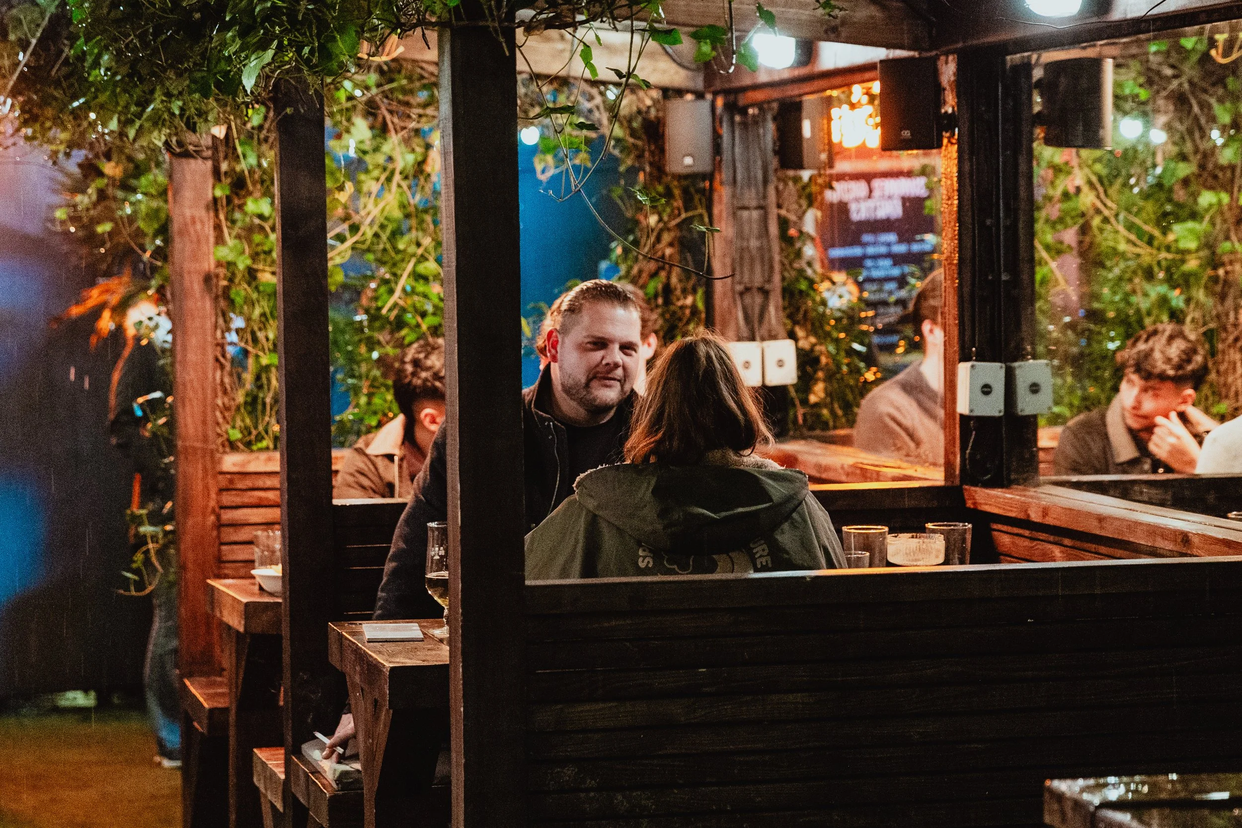People sitting at a table in a cozy, outdoor restaurant or bar, surrounded by greenery and warm lighting.