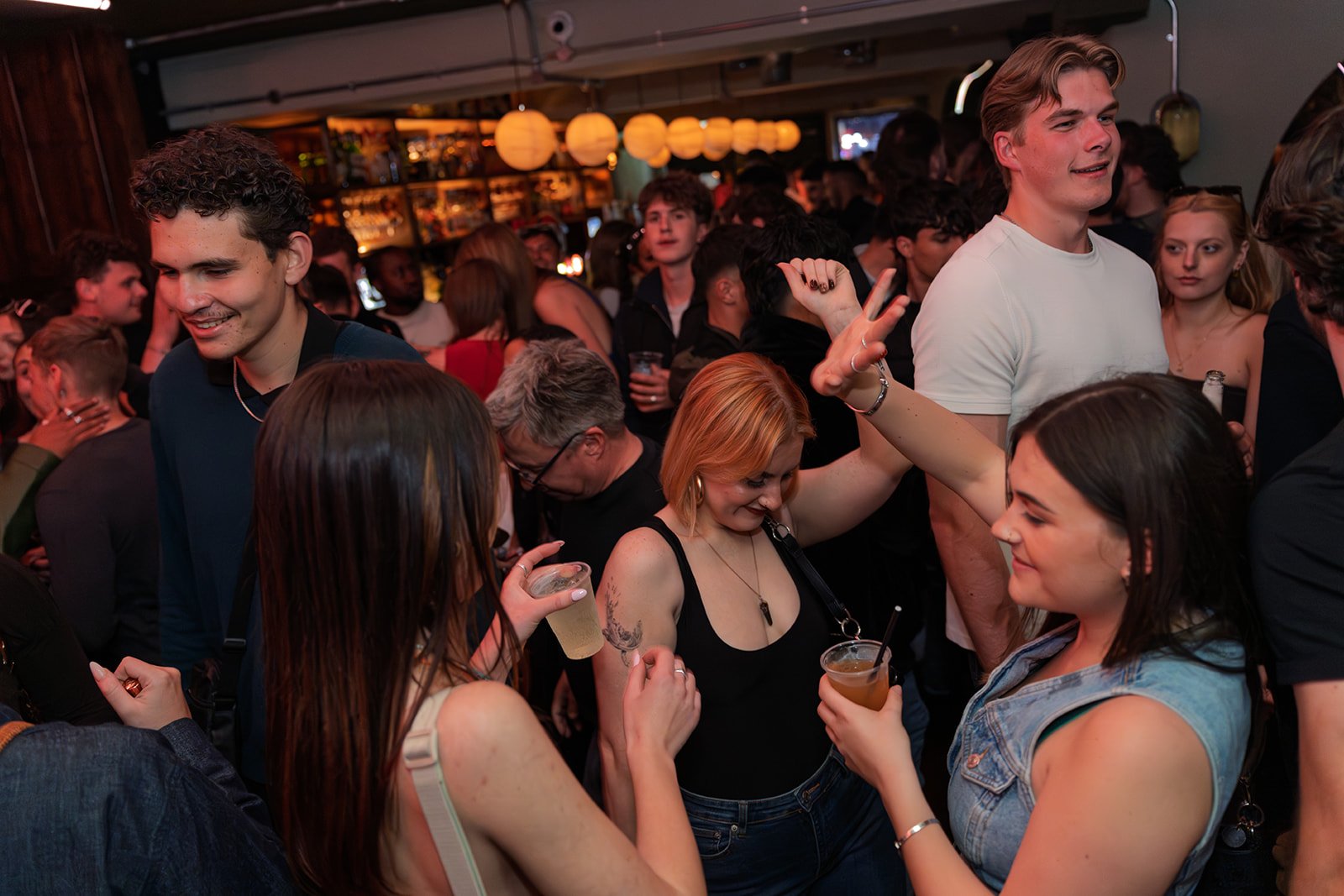 People dancing and socializing at a crowded bar or club, with warm lighting and tropical decor.