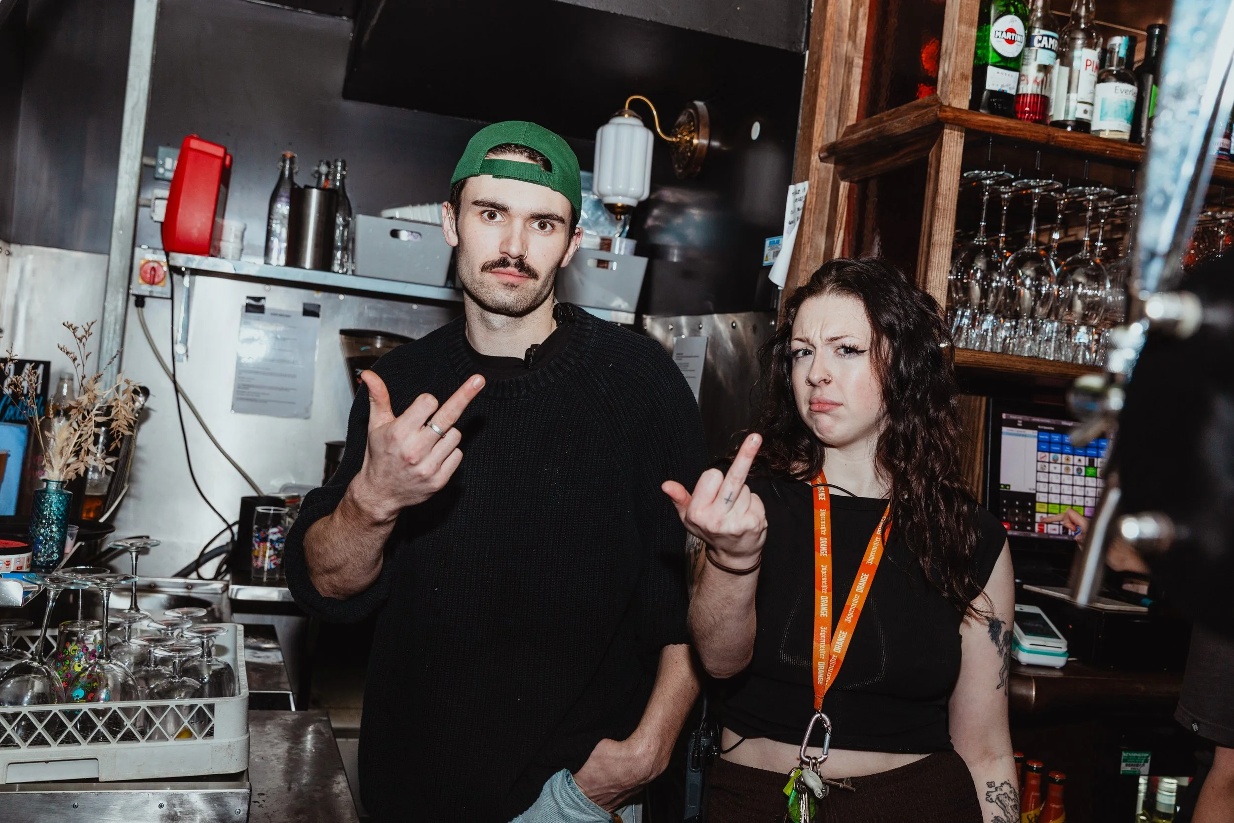 Two bartenders in a bar, both giving the middle finger and making facial expressions. The man wears a green cap backwards, the woman has curly dark hair and tattoos.