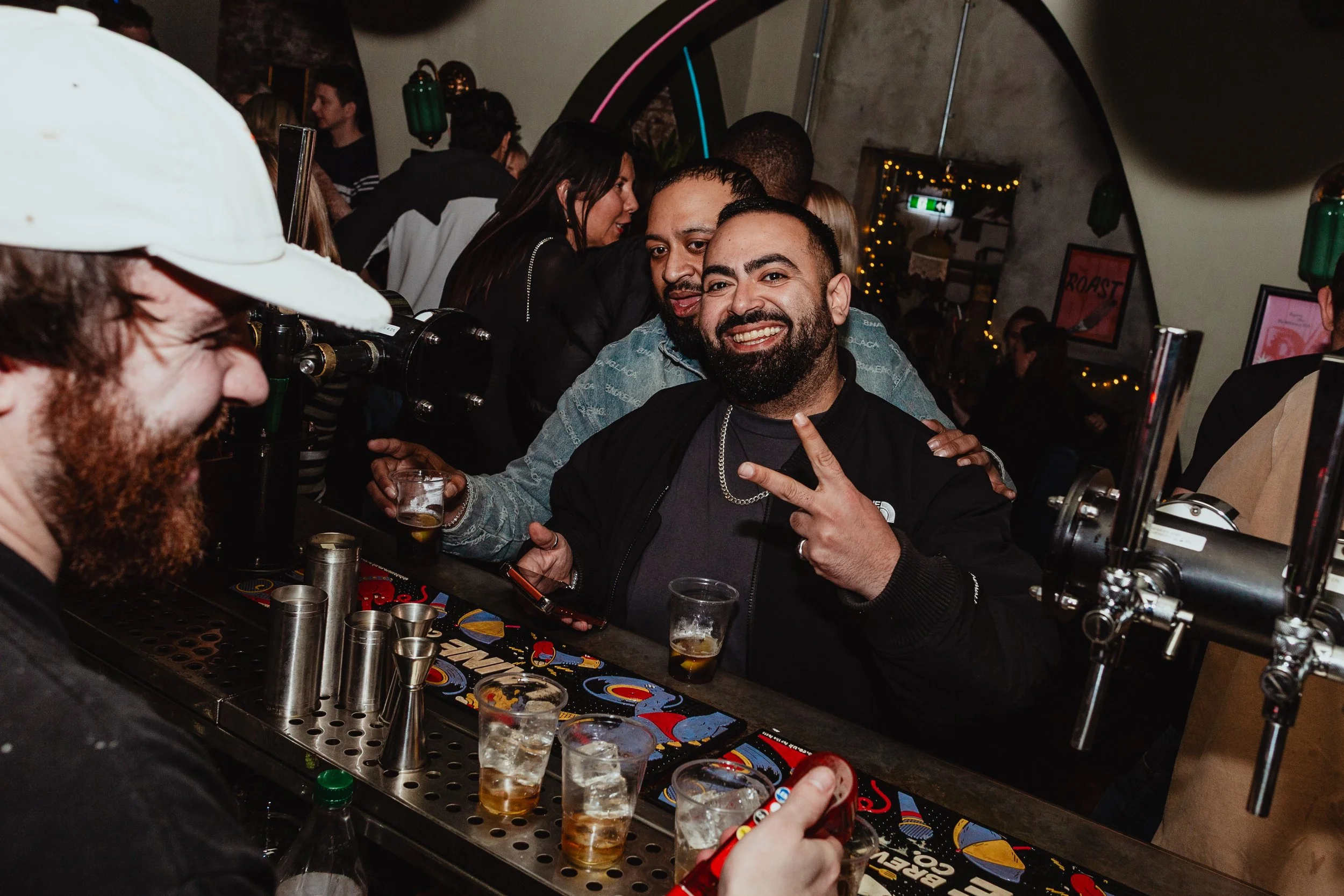 People enjoying drinks at a bar, with a man in the center flashing a peace sign and smiling.