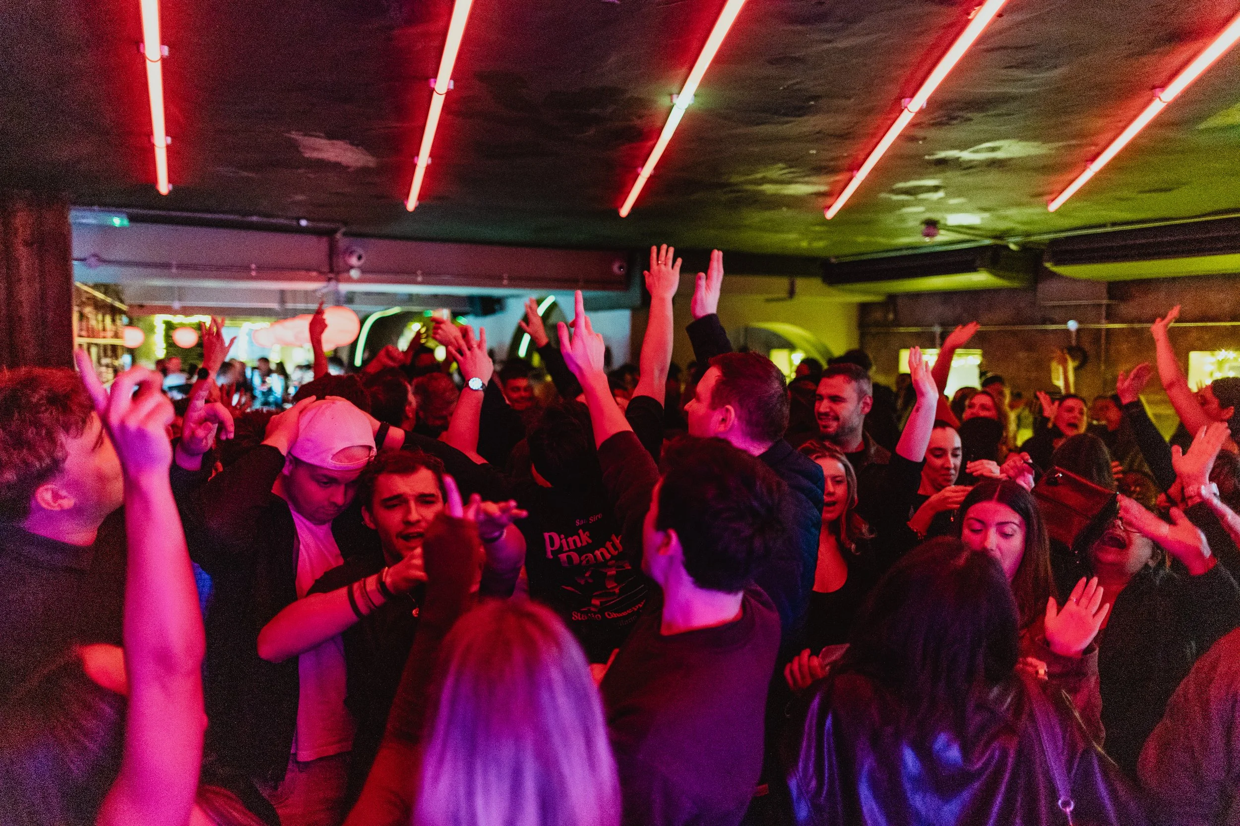 Crowd of people dancing and celebrating in a nightclub with colorful neon lighting and illuminated ceiling.