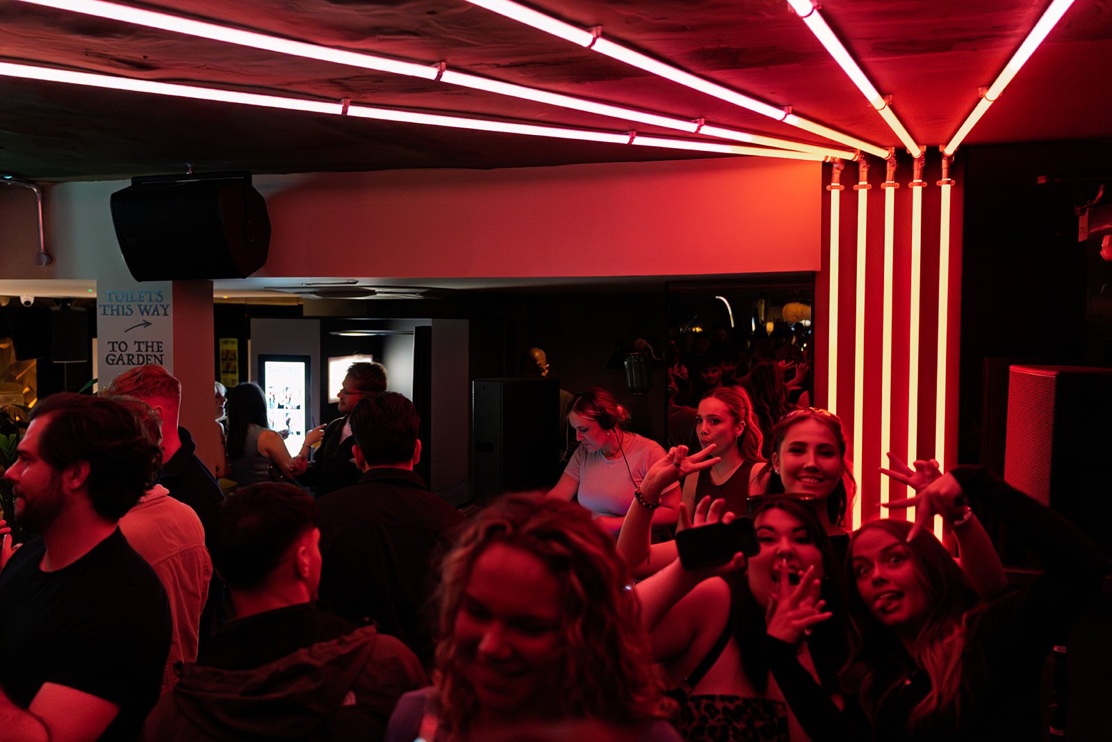 A group of people in a dimly lit nightclub with neon pink and white lights, some making peace signs and smiling.