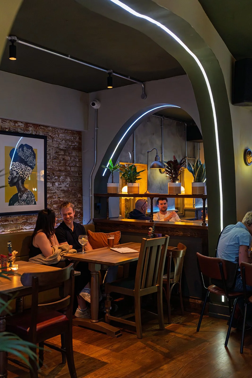 People sitting at tables in a cozy restaurant with brick walls, green arches with LED lights, and plants on a shelf.