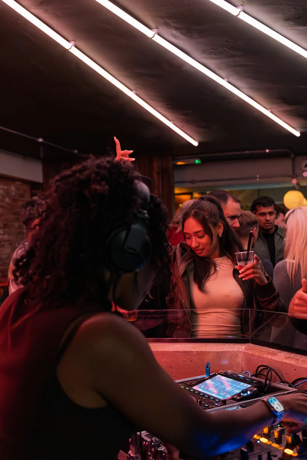 A female DJ wearing headphones performs at a nightclub with a crowd of people around her, some holding drinks, dancing and socializing under pink lighting and neon ceiling lights.