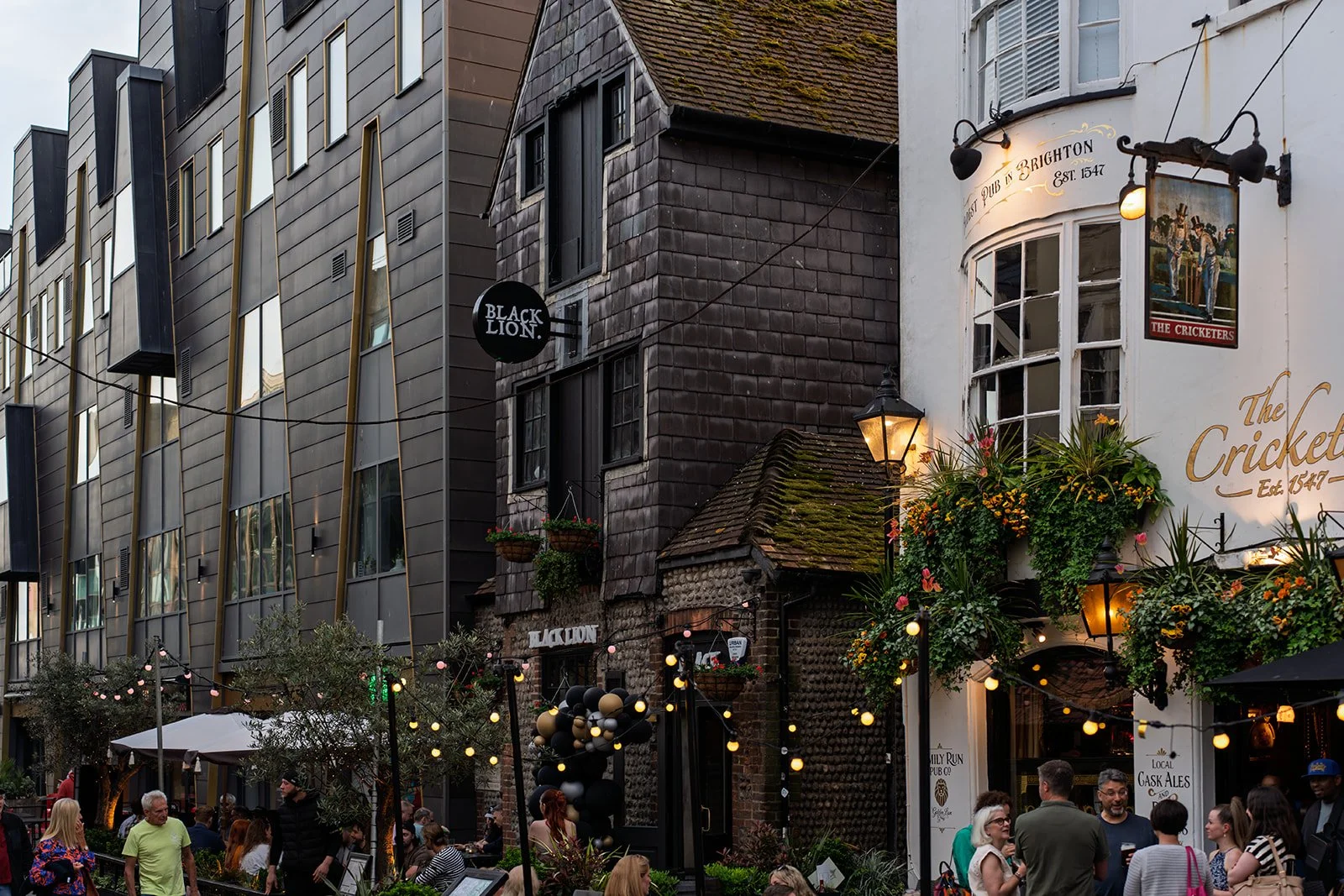 City street scene in the evening with people walking and gathering outside restaurants and bars, festive string lights hanging, and a mix of modern and historic buildings, including a pub with a white exterior and flower decorations.